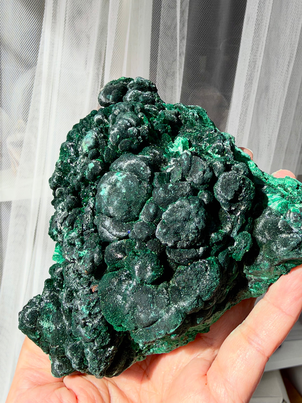 A hand holding a large, rough green malachite crystal cluster with a highly textured, bubble-like surface. The background features a white sheer curtain.