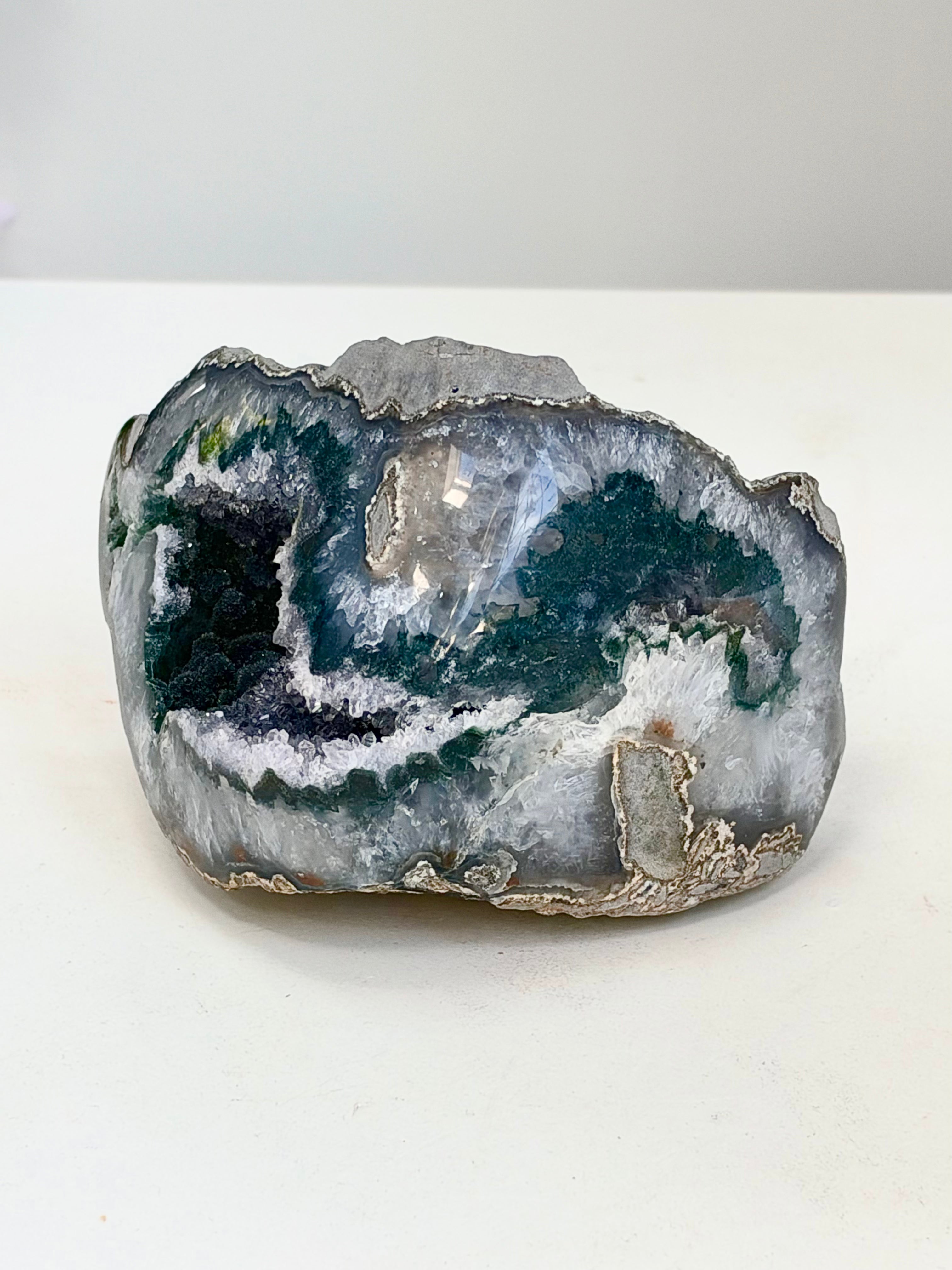 A raw geode specimen with a polished outer rind, featuring dark green, black, white, and gray crystalline interior formations on a plain white surface.