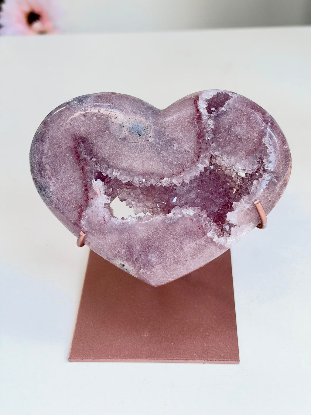 A heart-shaped pinkish-purple geode stone with white crystalline edges, displayed on a small rose-gold stand against a white background.
