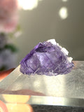 A raw purple fluorite crystal cluster with white quartz points, resting on a clear acrylic display block.
