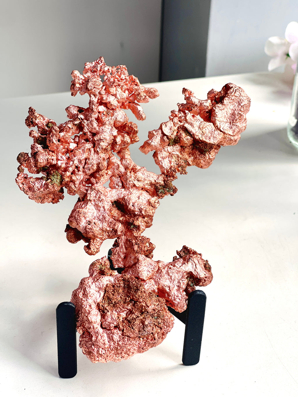 A metallic rose gold copper mineral specimen displayed on a black stand, set against a neutral indoor background.