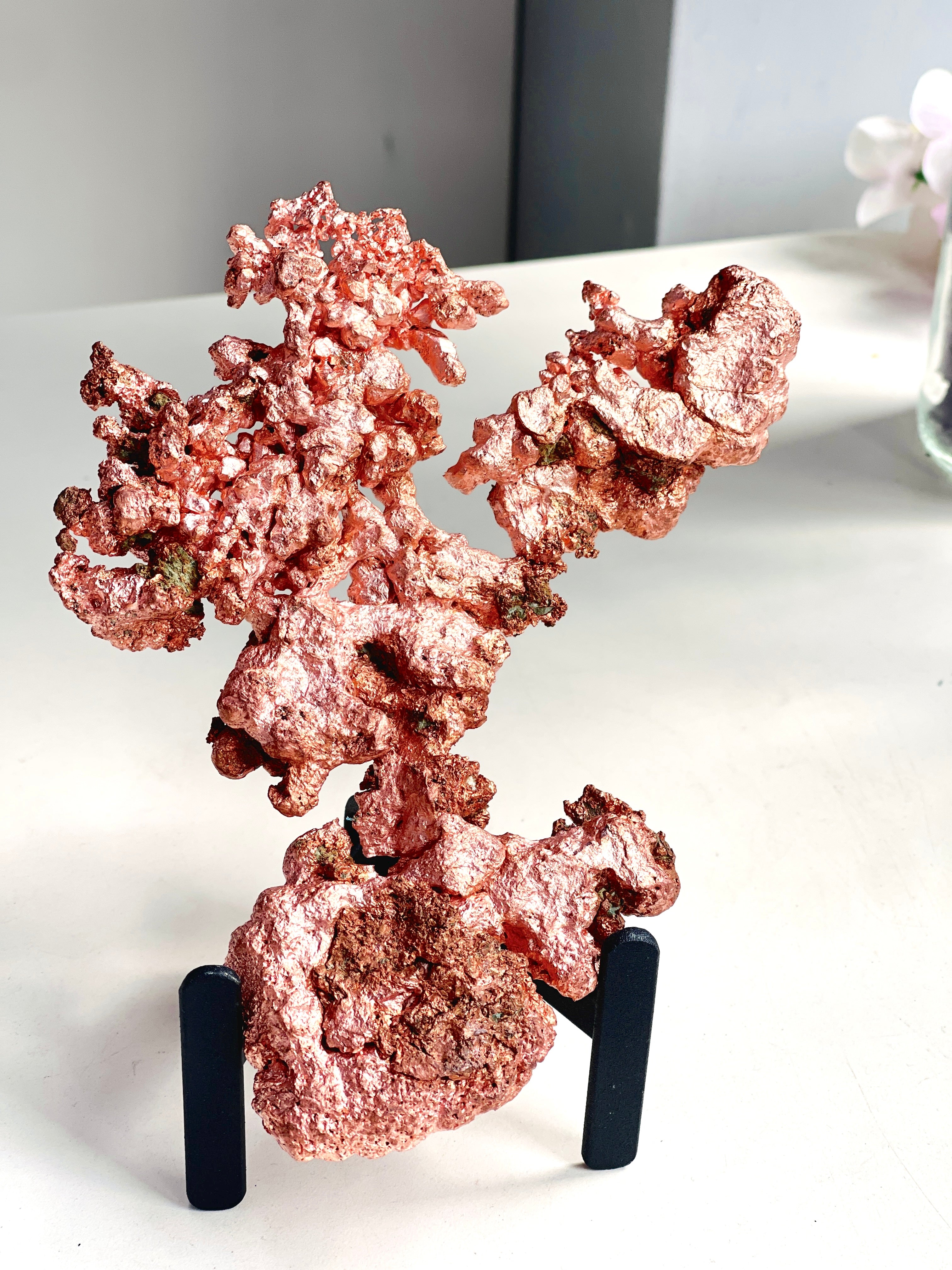A metallic rose gold copper mineral specimen displayed on a black stand, set against a neutral indoor background.