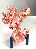 A metallic copper-colored mineral specimen or decorative sculpture with a rough, textured surface, displayed on a small dark blue stand against a plain light gray and white background.