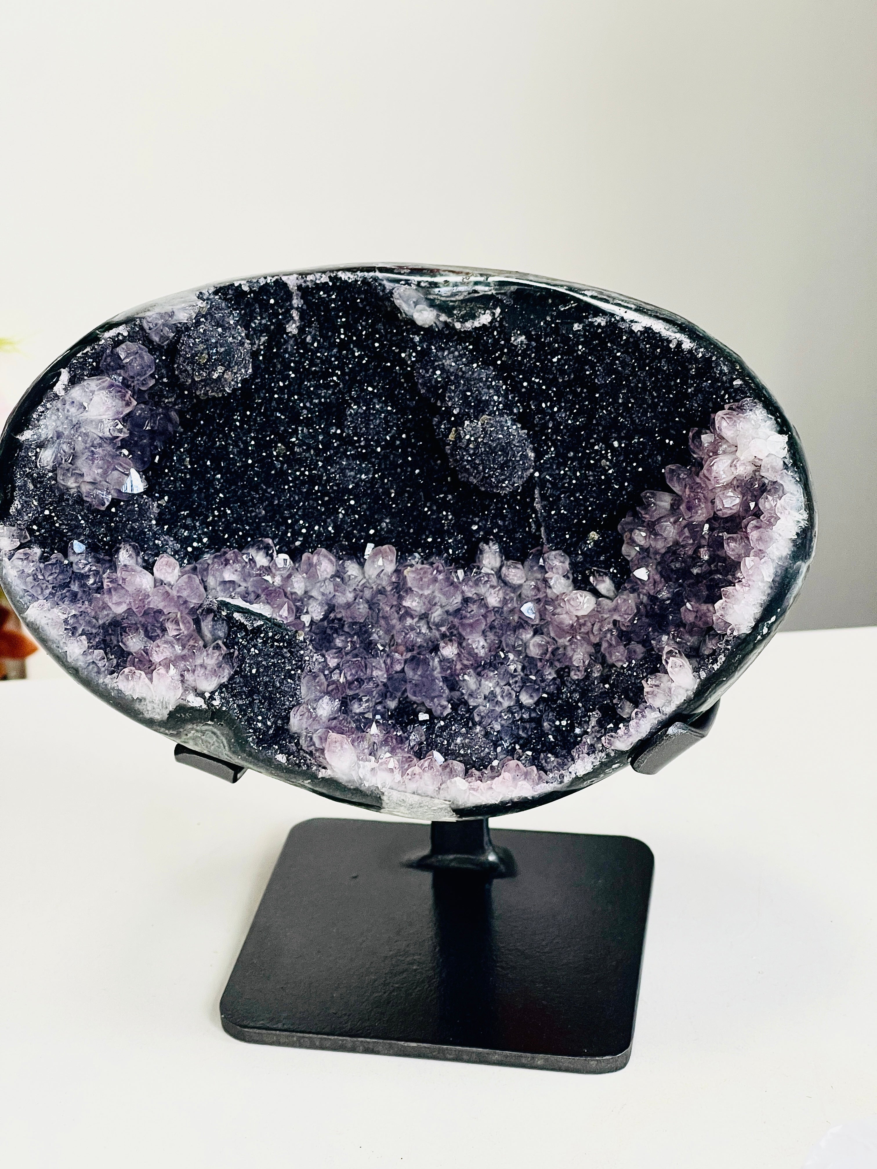 A polished amethyst geode specimen with a black cavity and purple crystal formations, displayed on a black metal stand against a plain light background.