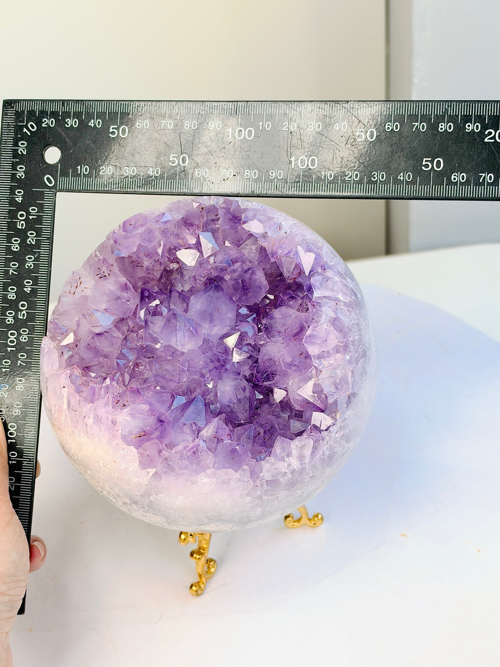 A purple amethyst geode sphere on a small gold stand, shown with a metal ruler for scale against a plain indoor background.