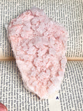 A pale pink halite crystal cluster or raw gemstone with rough cuboid facets, photographed from above on top of printed book pages.