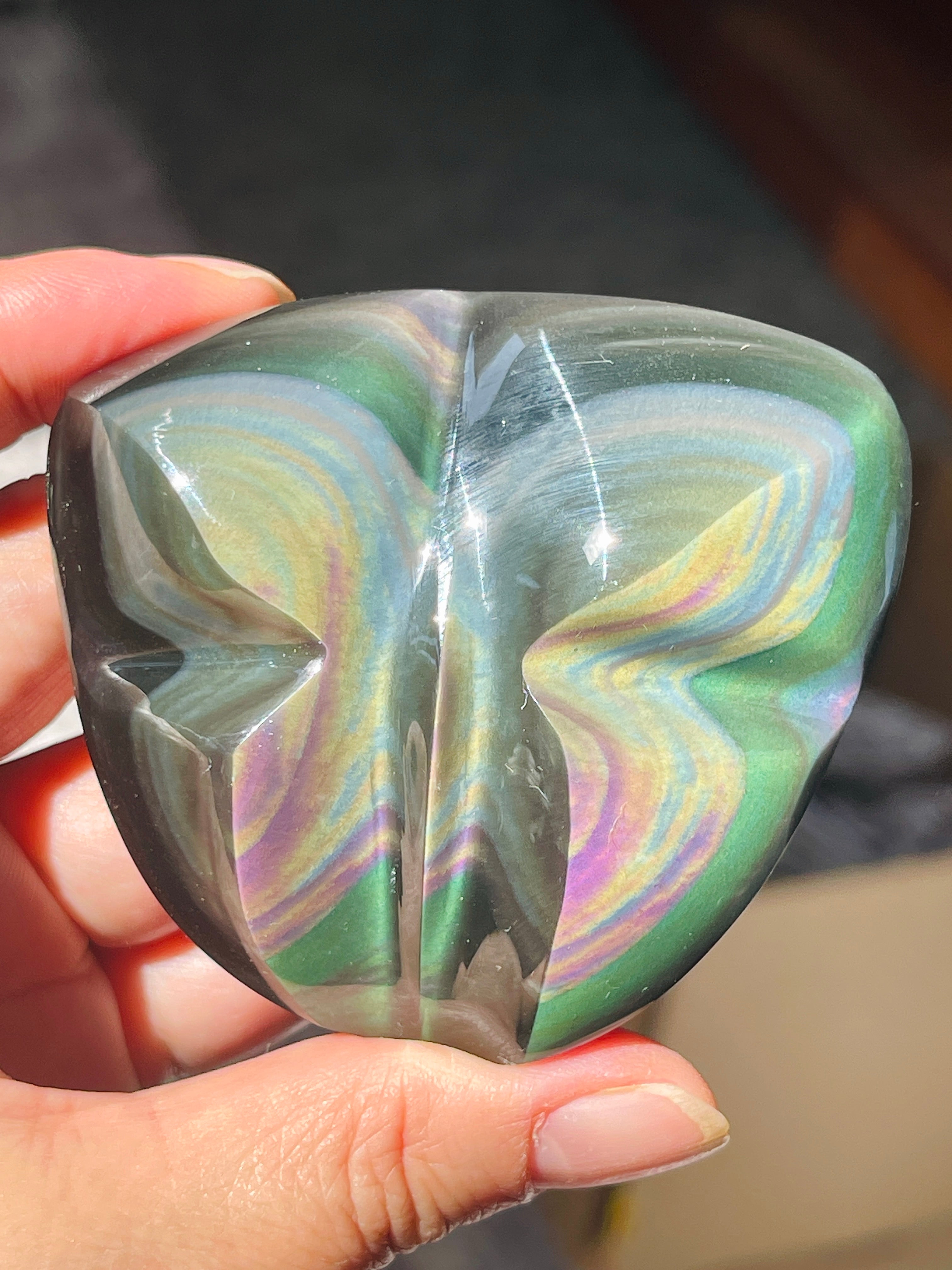 A butterfly-shaped iridescent stone held in a hand, showing swirling green, blue, yellow, and purple colors on a dark background.
