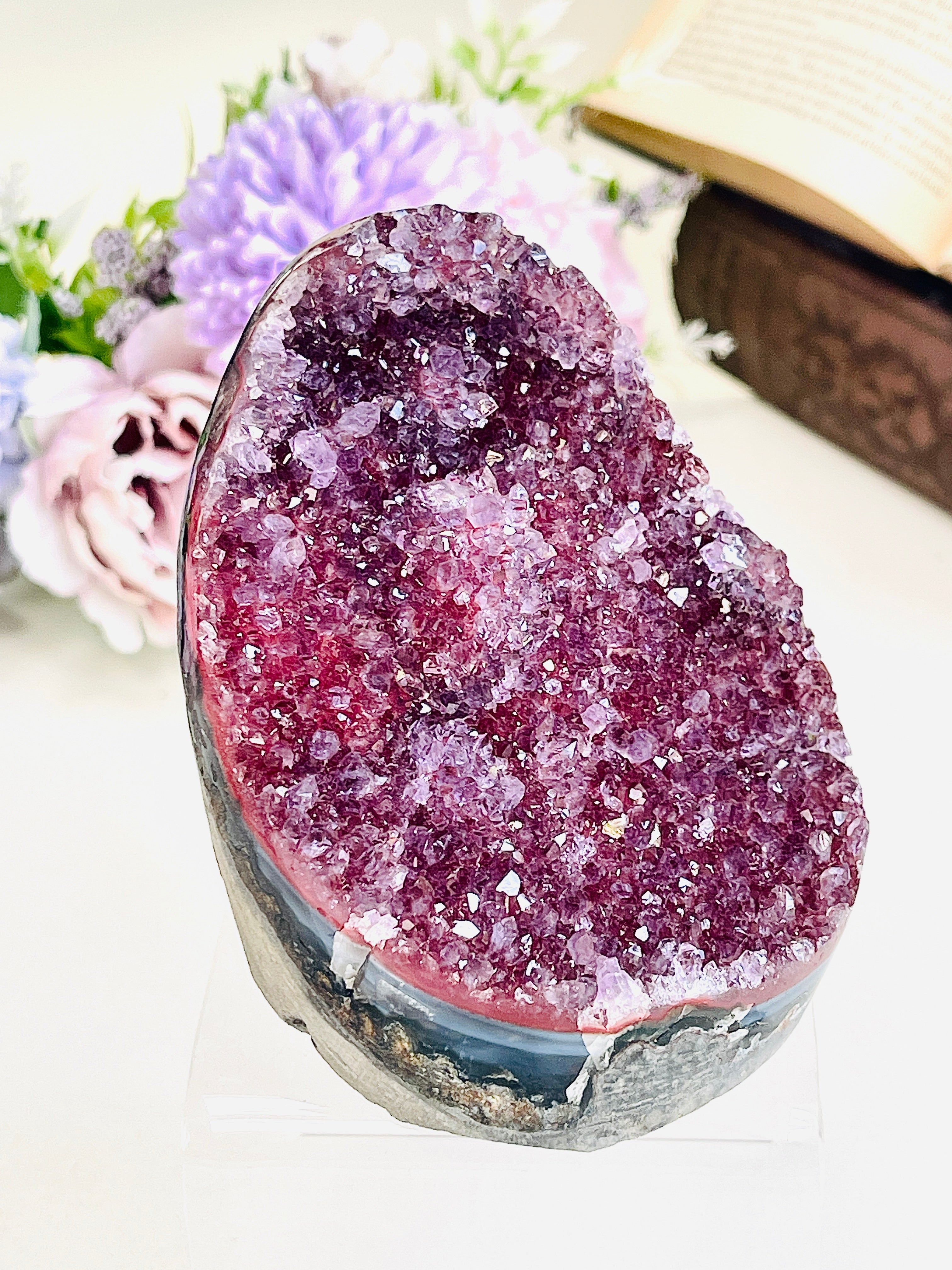 A raw amethyst geode specimen with a sparkling purple crystal cavity and a dark natural base, displayed on a clear acrylic stand against a softly blurred background of flowers and an open book.