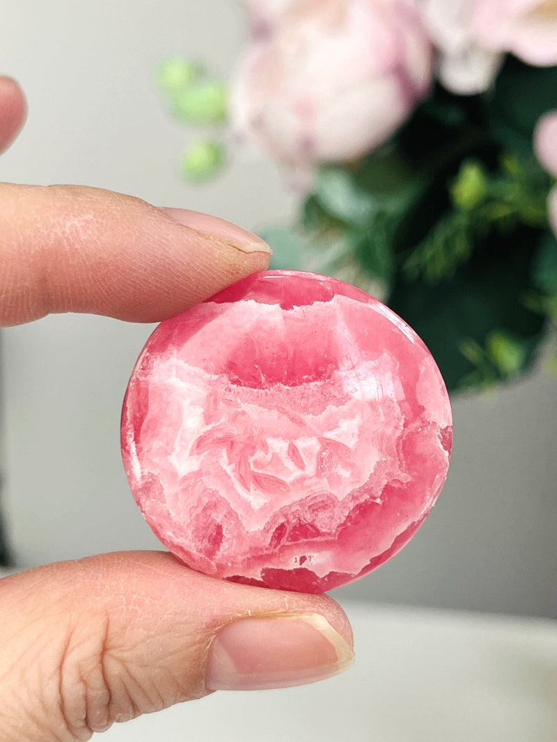 a round pink rhodochrosite crystal crystal with pale pink patterns hold by two fingers , with a some flower in a white background 