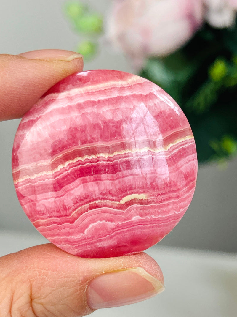 Pink and white striped rhodochrosite stone held between fingers with a blurred floral background